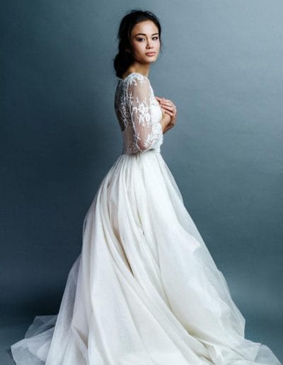 Eine Braut in einem weißen Hochzeitskleid posiert vor grauem Hintergrund.